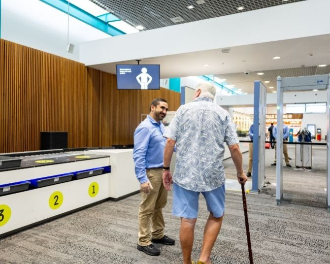 Airport security checkpoint with an elderly man using a cane approaching the screening area, assisted by a security officer near numbered bins and instructional signage.