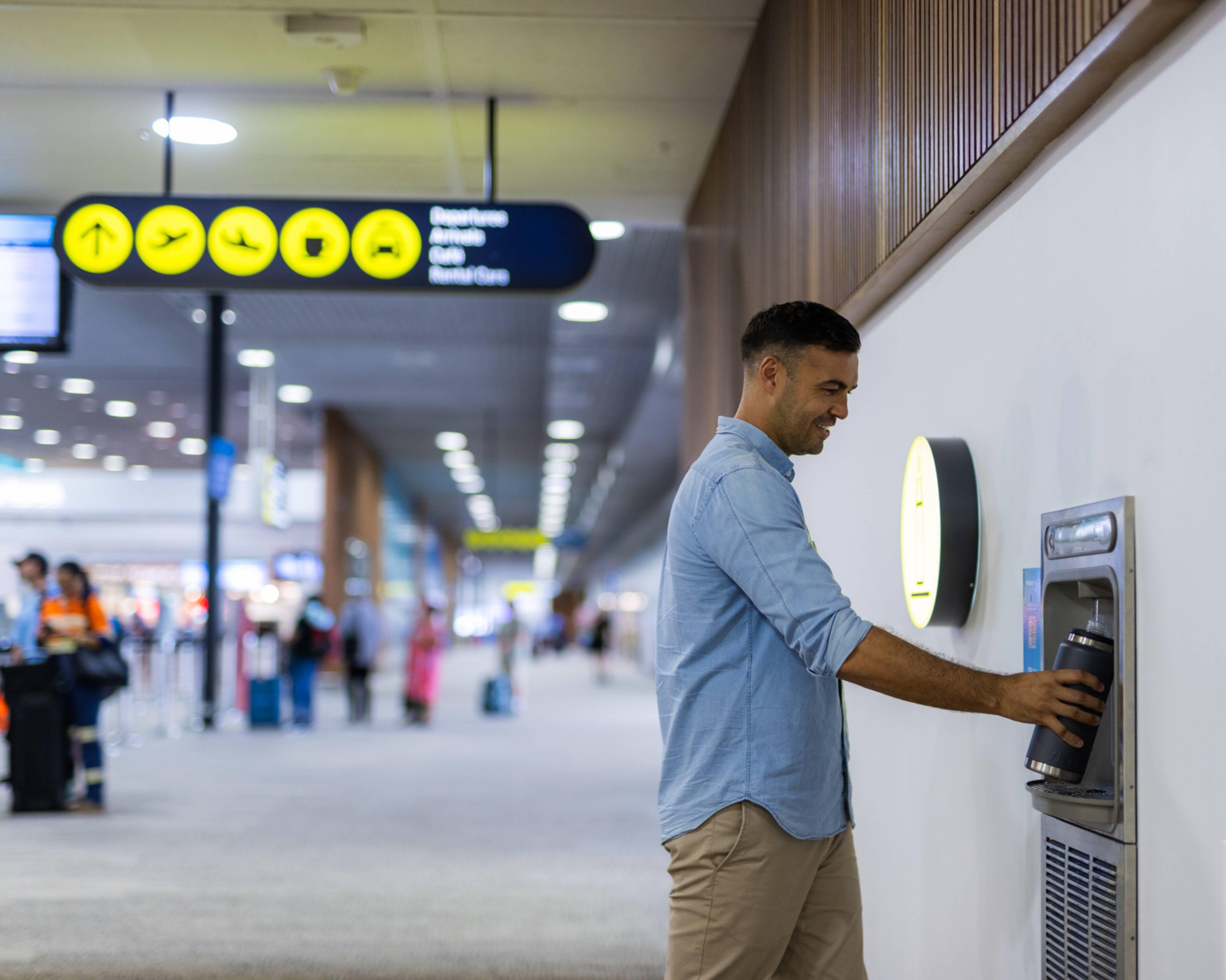 Traveler refills a water bottle at an airport hydration station, with directional signage and terminal activity visible in the background.