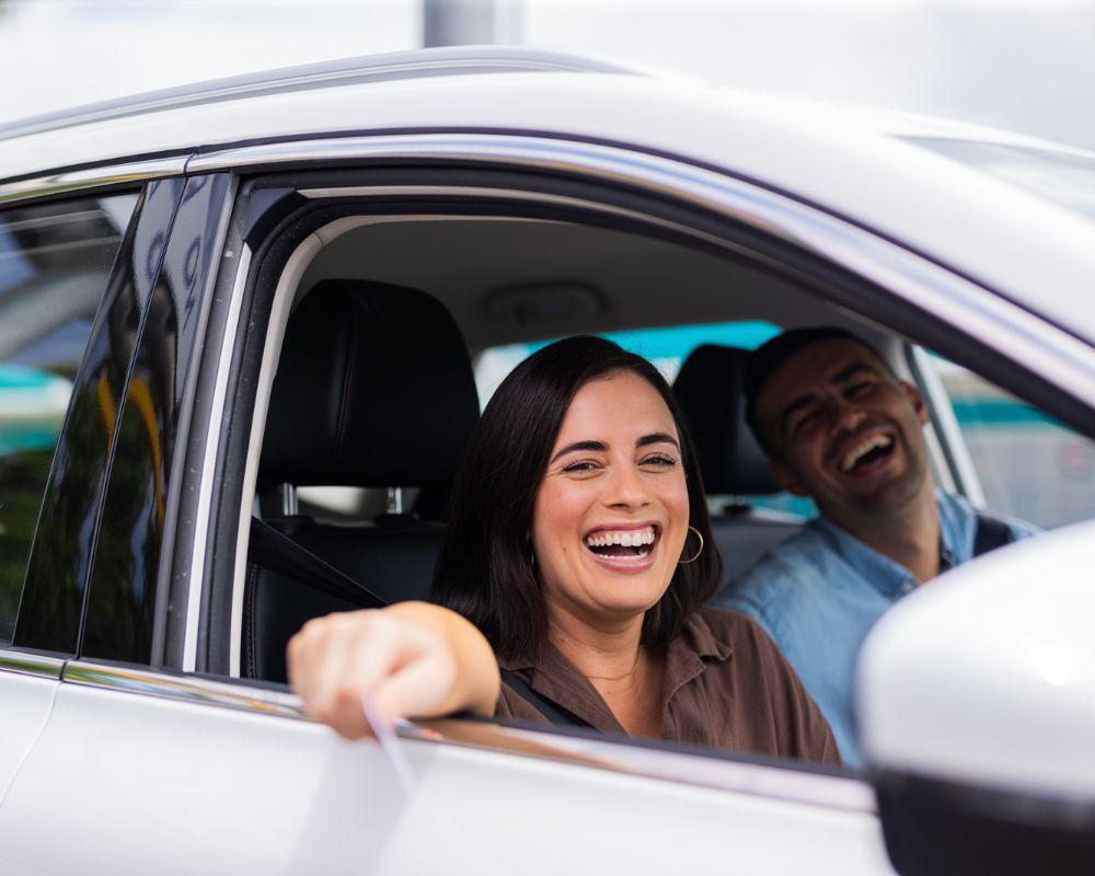 Two people sit inside a white car with laughter, with the front passenger extending an arm out the window holding a document.