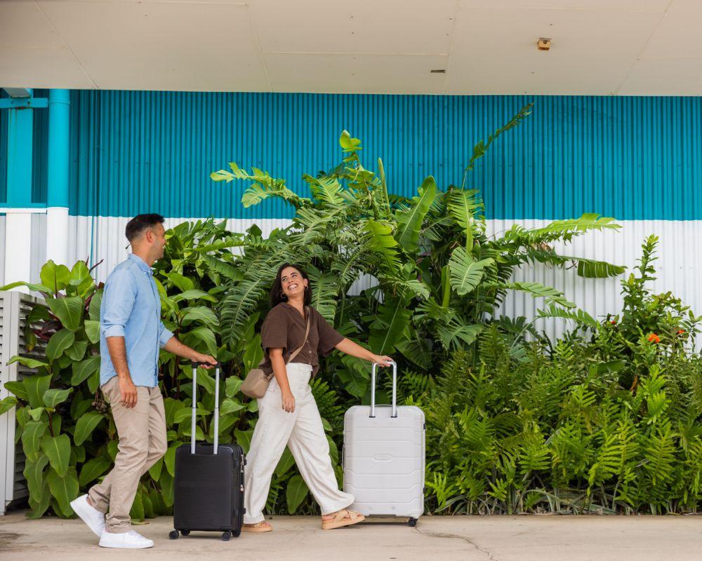 Two travelers walk with rolling suitcases in front of a blue and white building, surrounded by lush green plants.